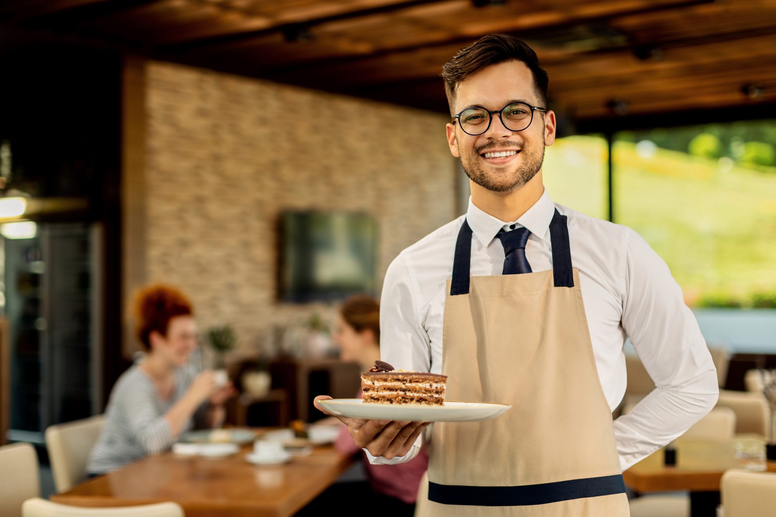 Happy waiter with slice of cake standing in a cafe and looking at camera. There are people in the background.